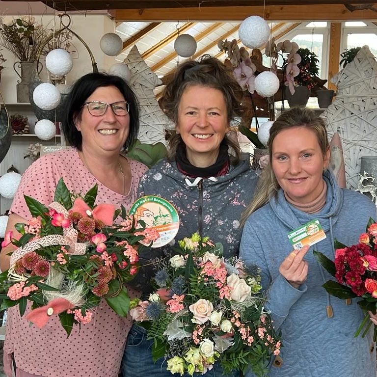 Drei Frauen lächeln, halten Blumengestecke und eine Grußkarte in einem Blumenladen.